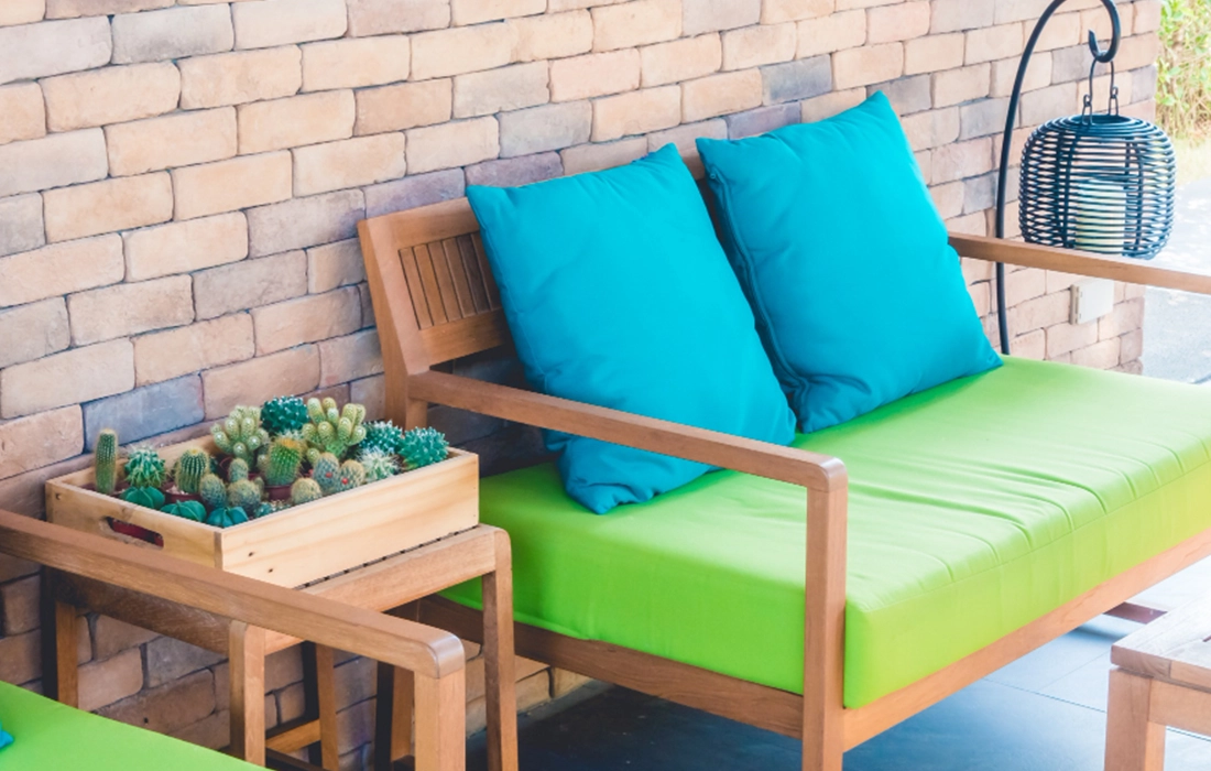 Wooden bench with bright green cushions and two blue pillows beside a small table holding a wooden box of various cacti, set against a brick wall.