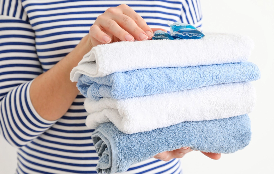 A person in a striped shirt holds a stack of neatly folded blue and white towels with a laundry detergent pod on top.