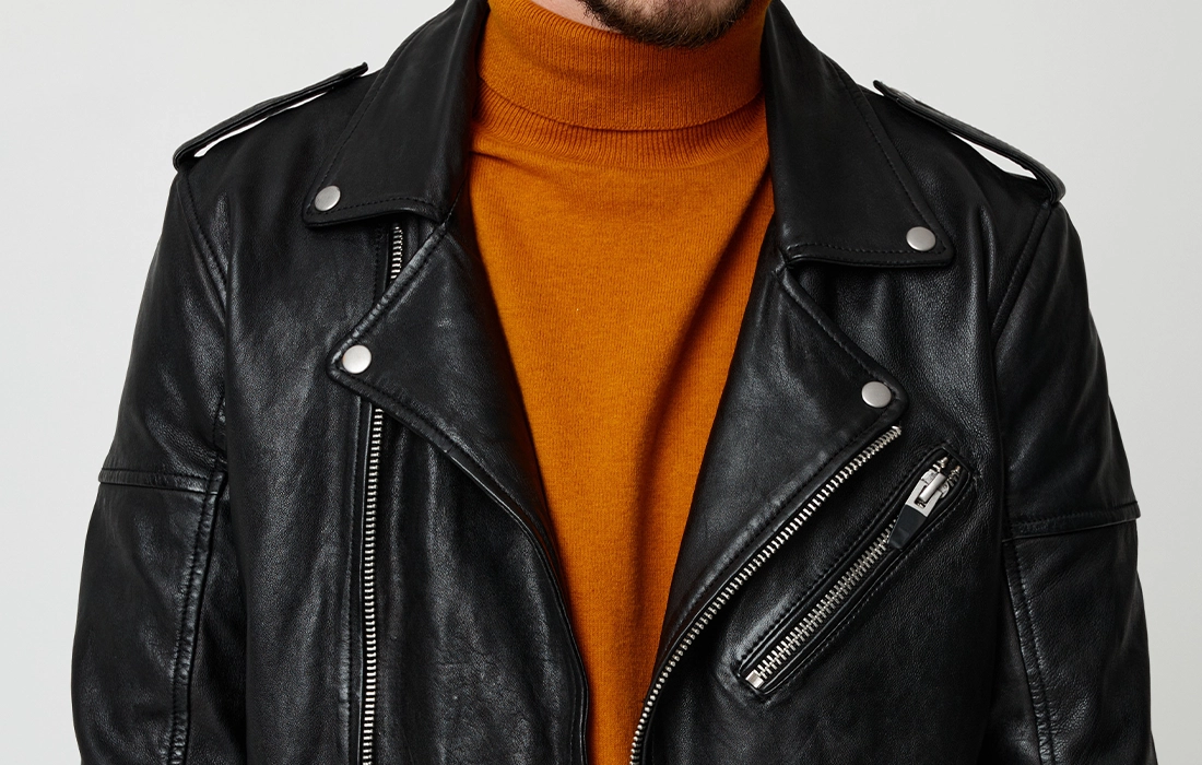 A person wearing a black leather biker jacket over an orange turtleneck sweater against a plain background.