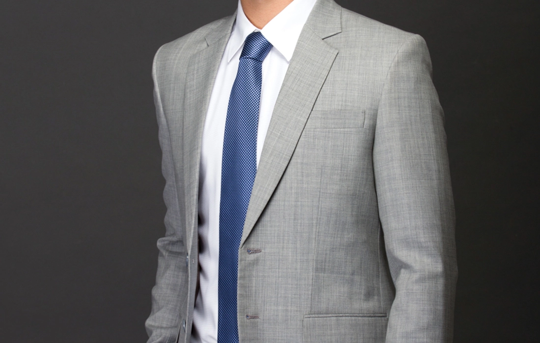A person wearing a light gray suit, white dress shirt, and blue patterned tie against a dark background.