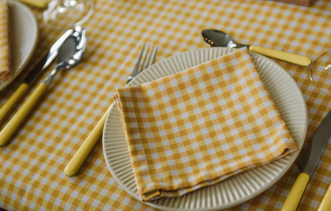 A plate with a yellow and white checkered napkin is set on a matching tablecloth, accompanied by yellow-handled cutlery.