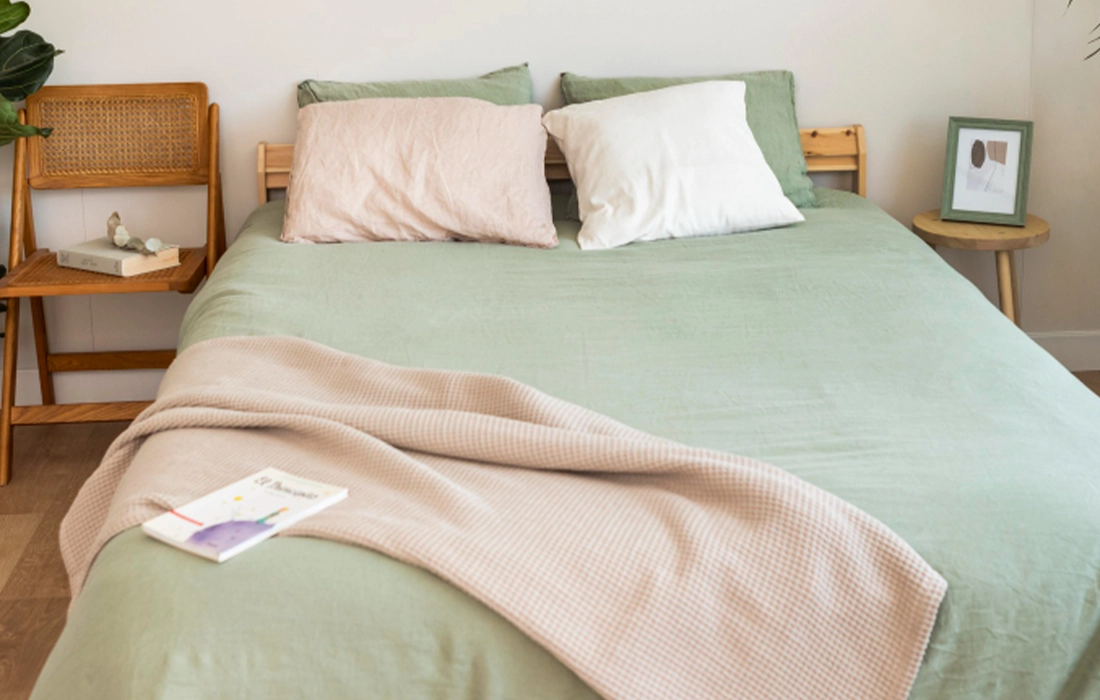 A neatly made bed with green bedding, two pillows, and a beige blanket. A book rests on the bed, and a chair and framed picture sit beside the bed.