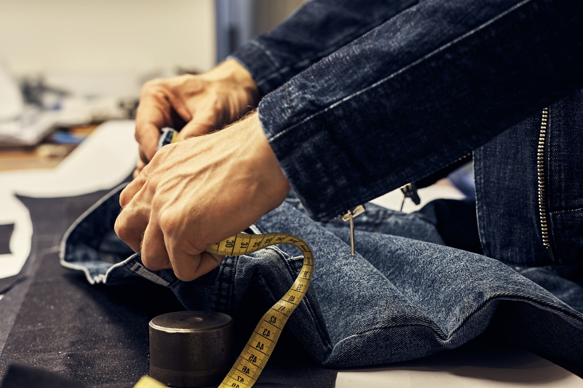 Close-up of a person measuring denim fabric with a yellow measuring tape, wearing a dark denim jacket, in a tailoring or textile workspace.