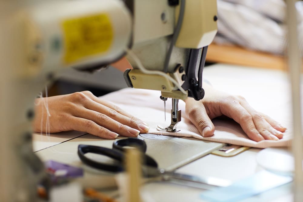Close-up of hands guiding fabric through a sewing machine, with scissors and other sewing tools nearby on the table.