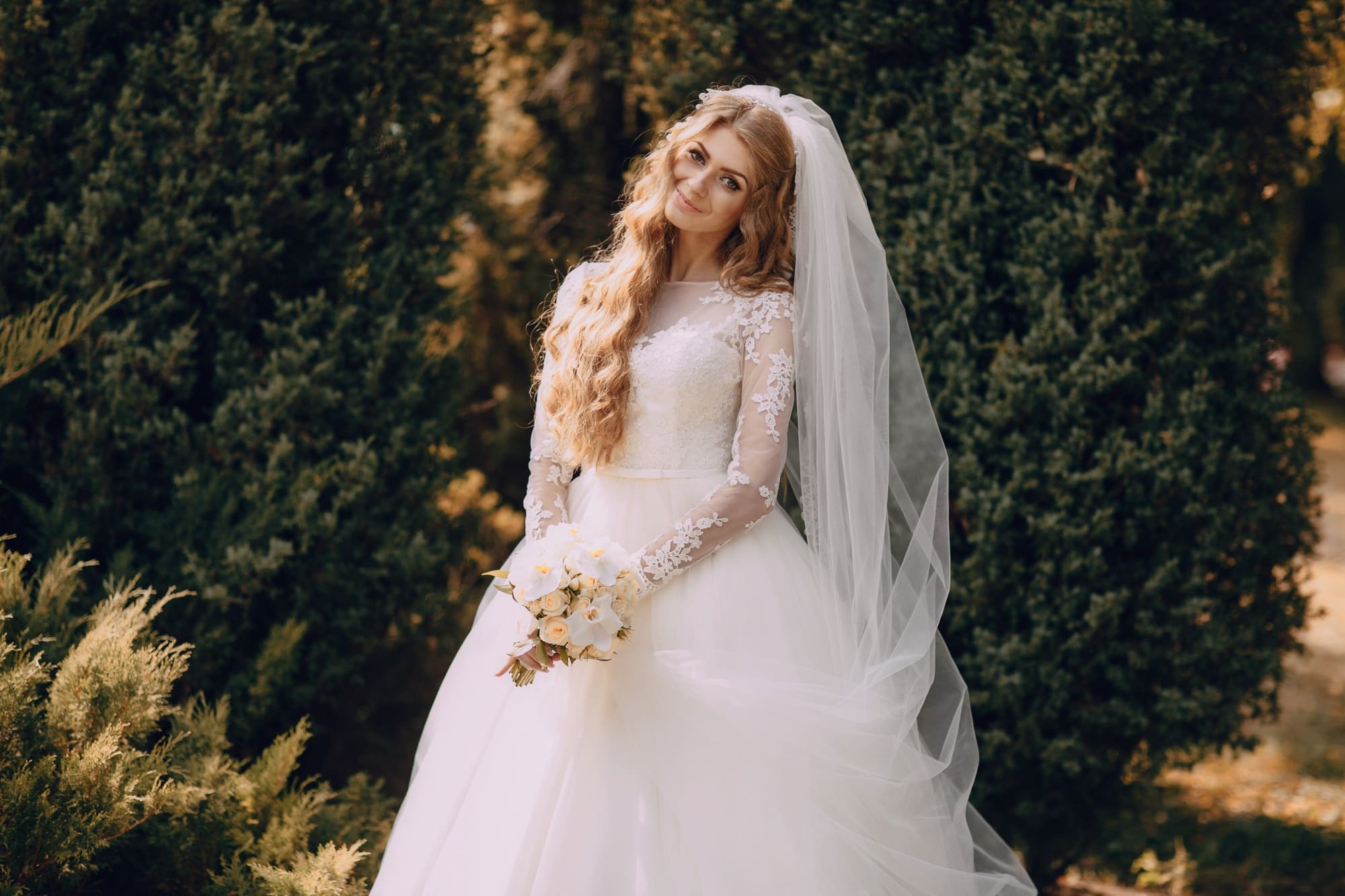 A woman in a white wedding dress and veil stands outdoors holding a bouquet, with greenery in the background.