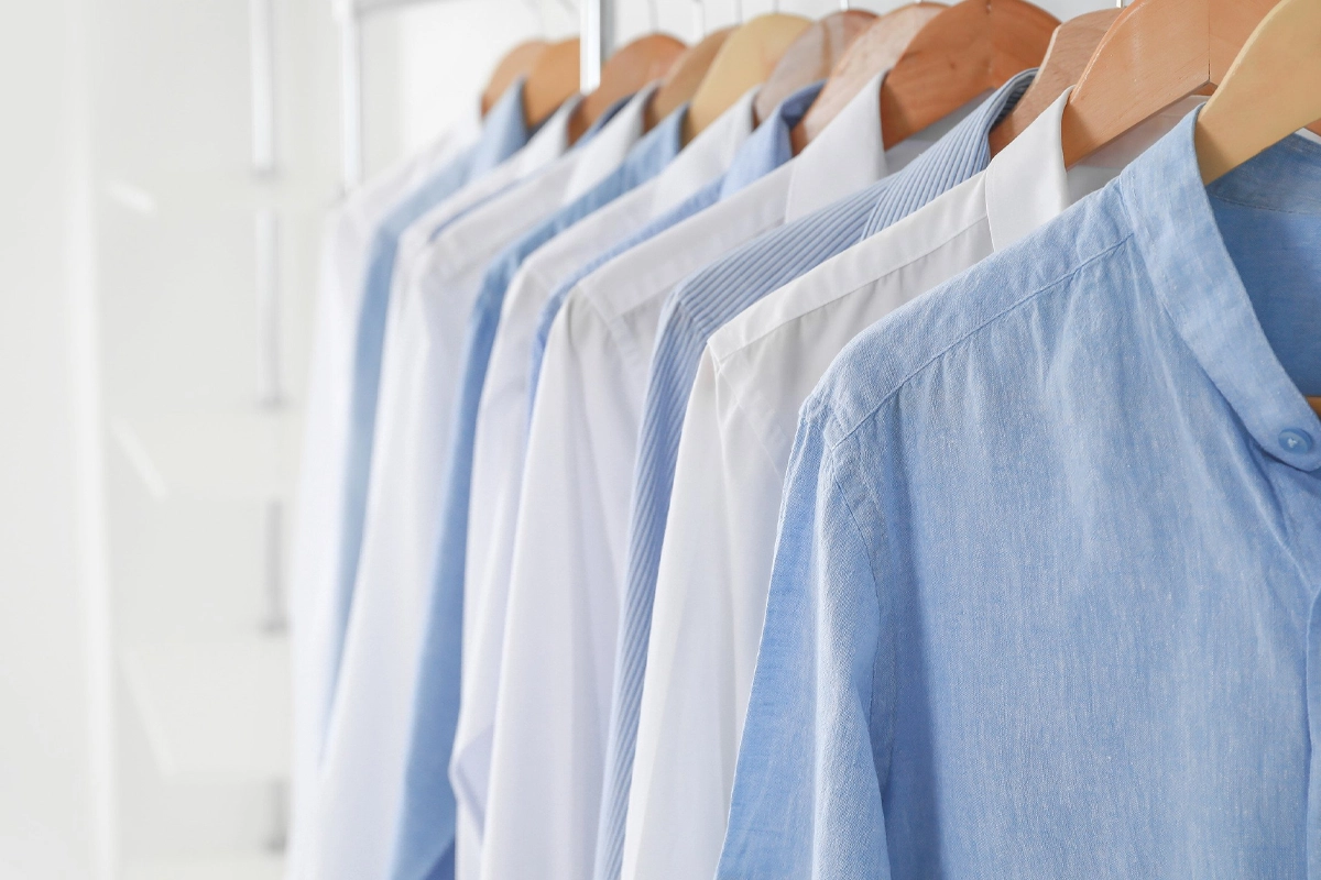 A row of light blue and white button-up shirts hangs neatly on wooden hangers in a well-lit closet.