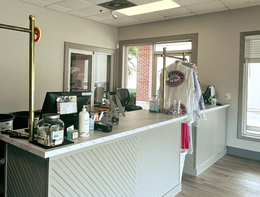 A dry cleaning shop counter with a computer, cash jar, hand sanitizer, and freshly cleaned clothes hanging on a rack.