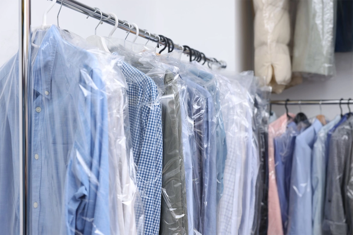 Clothes hang on a rack, each covered in clear plastic garment bags, including shirts in various colors and patterns, in a dry cleaning facility.