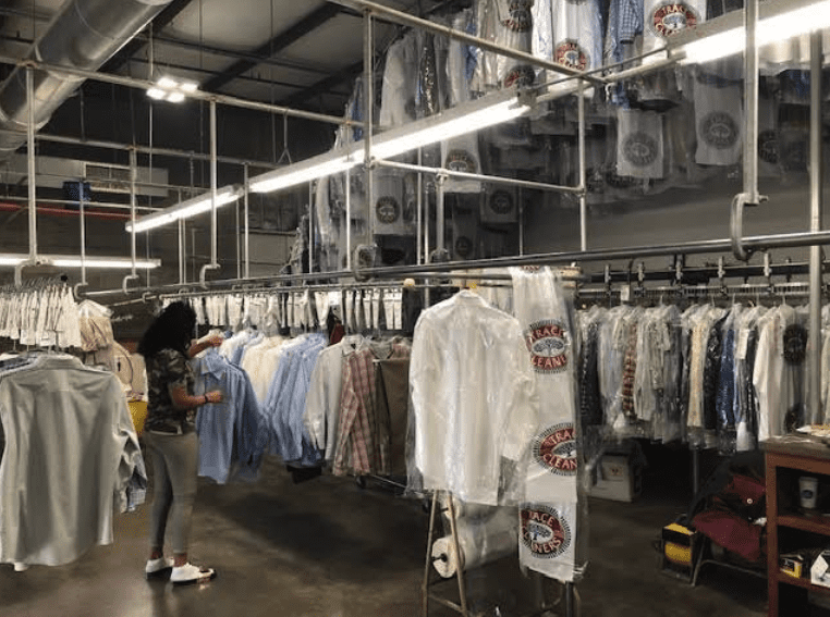 A woman stands in a dry cleaning facility, sorting through racks of washed and plastic-wrapped clothes under fluorescent lights.