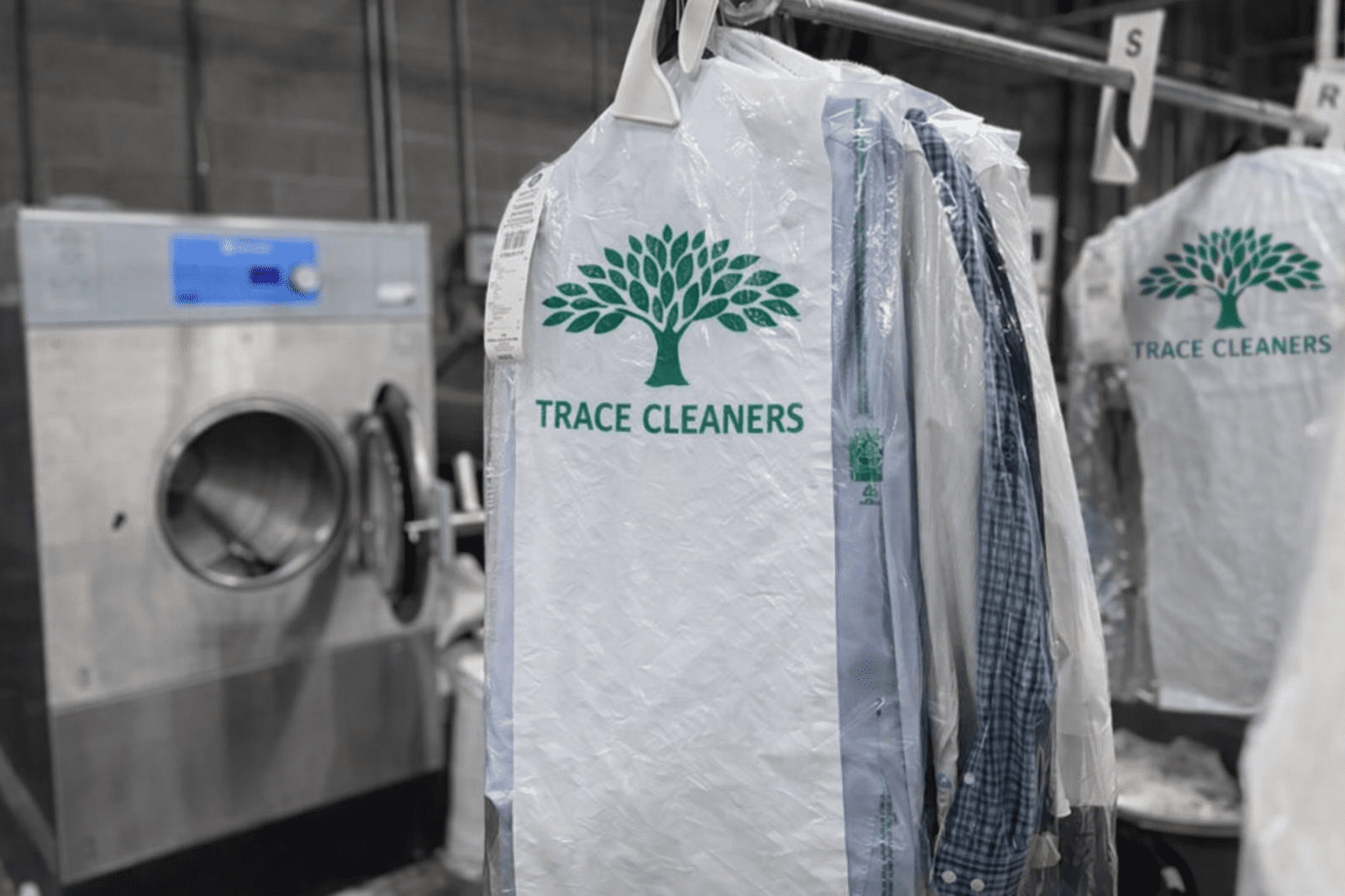 Clothes on hangers covered in Trace Cleaners plastic garment bags are near an industrial washing machine in a laundry facility.