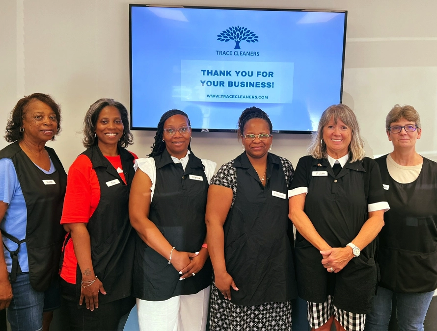 Six women wearing black aprons stand in front of a screen displaying a Trace Cleaners thank you message.