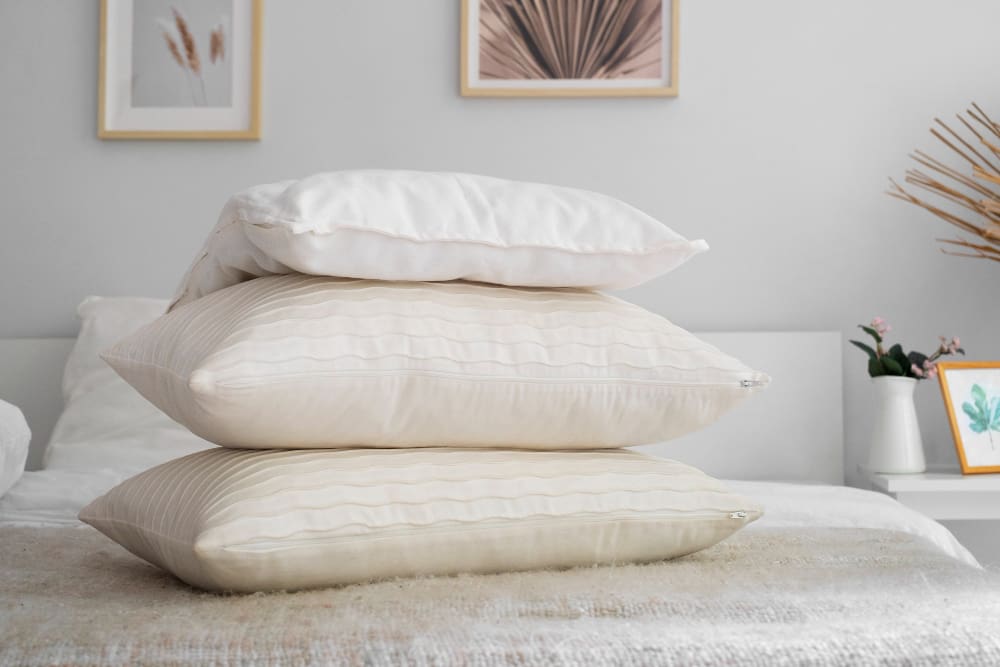 Three pillows are stacked on a bed in a minimalist bedroom with light-colored decor and framed artwork on the wall.