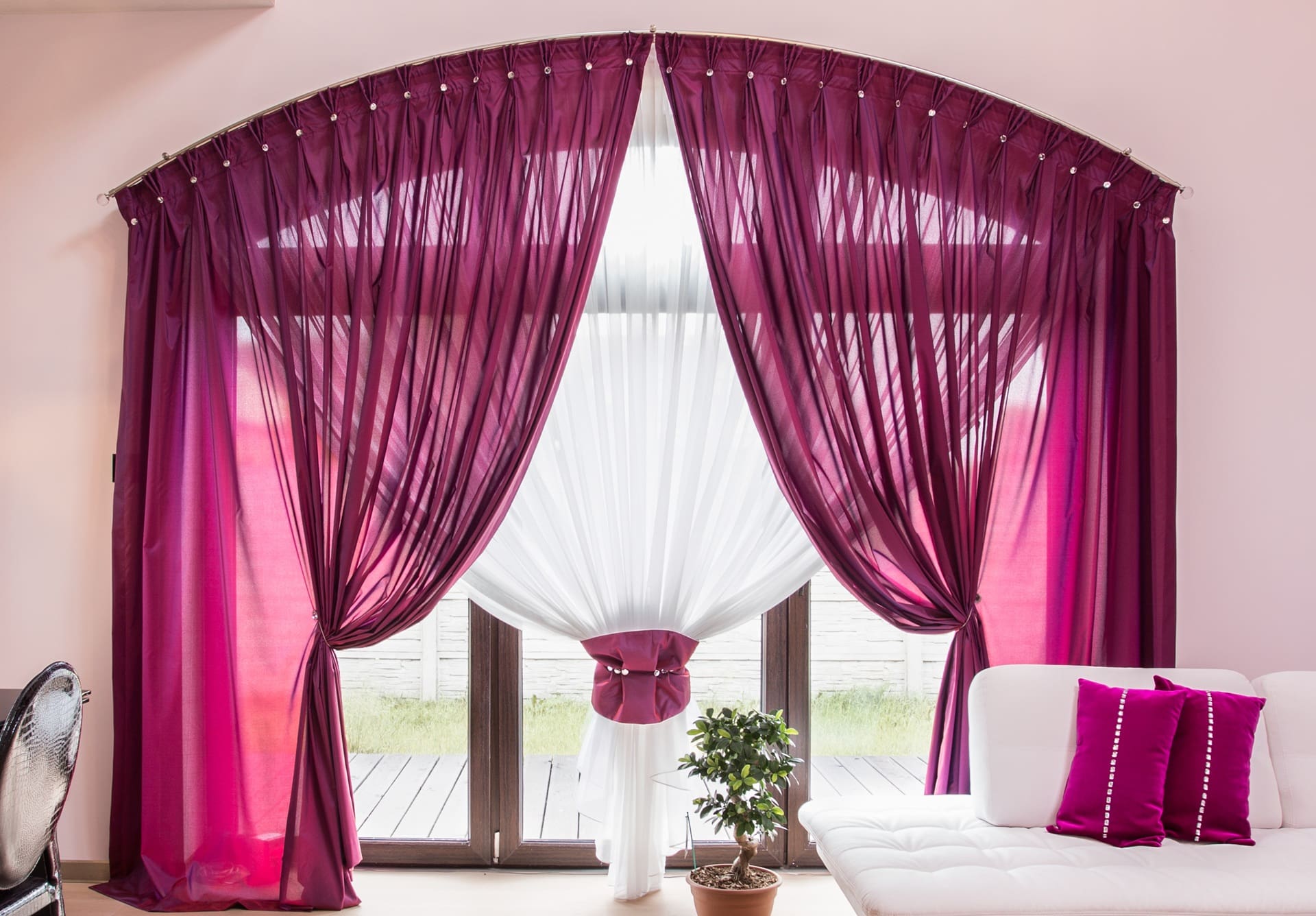 Large window with sheer white curtains and dark pink drapes tied back, letting in natural light; a potted plant sits in front and a white sofa with pink pillows is on the right.