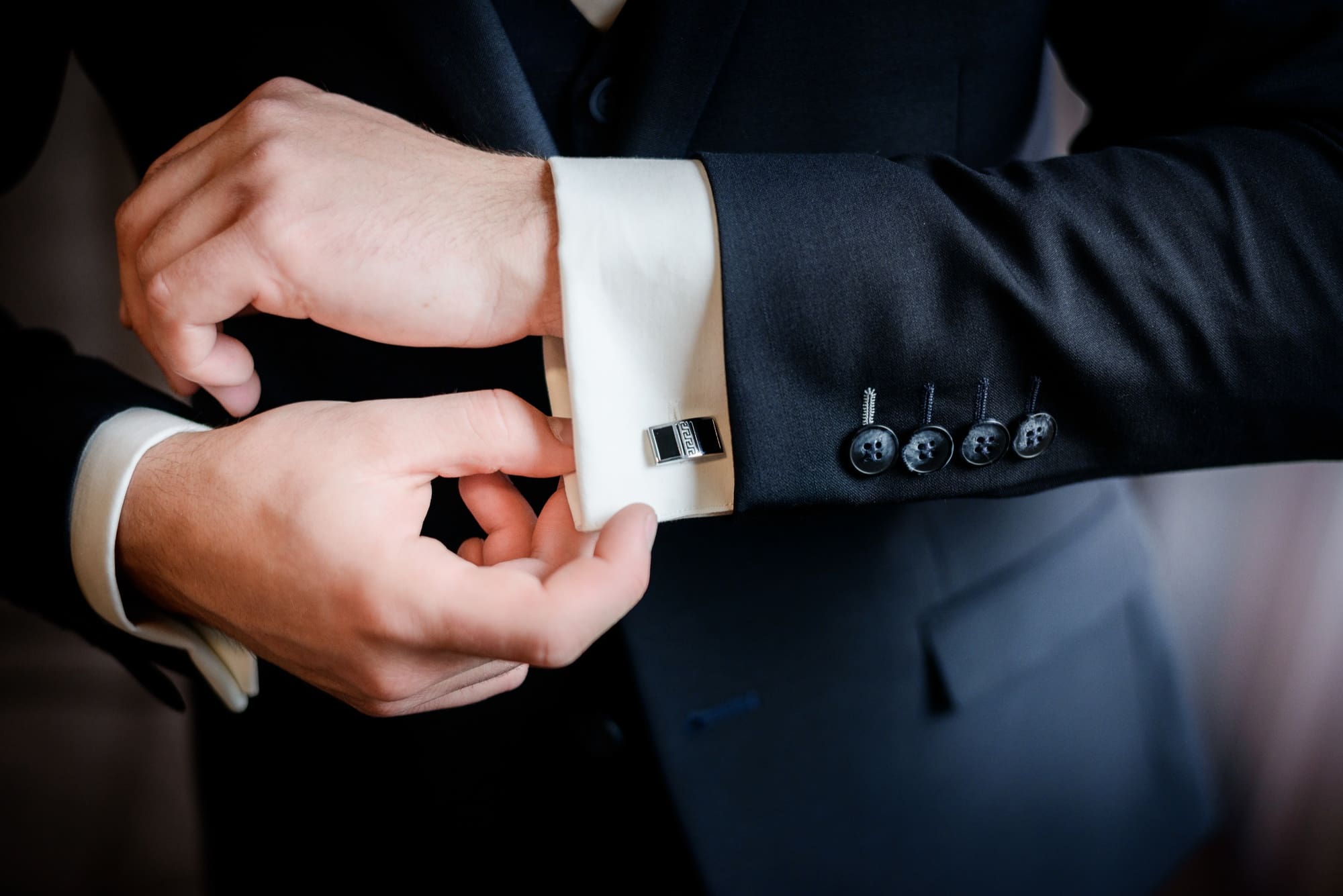 A person in a dark suit adjusts a silver cufflink on their white shirt sleeve, with four buttons visible on the jacket&rsquo;s cuff.