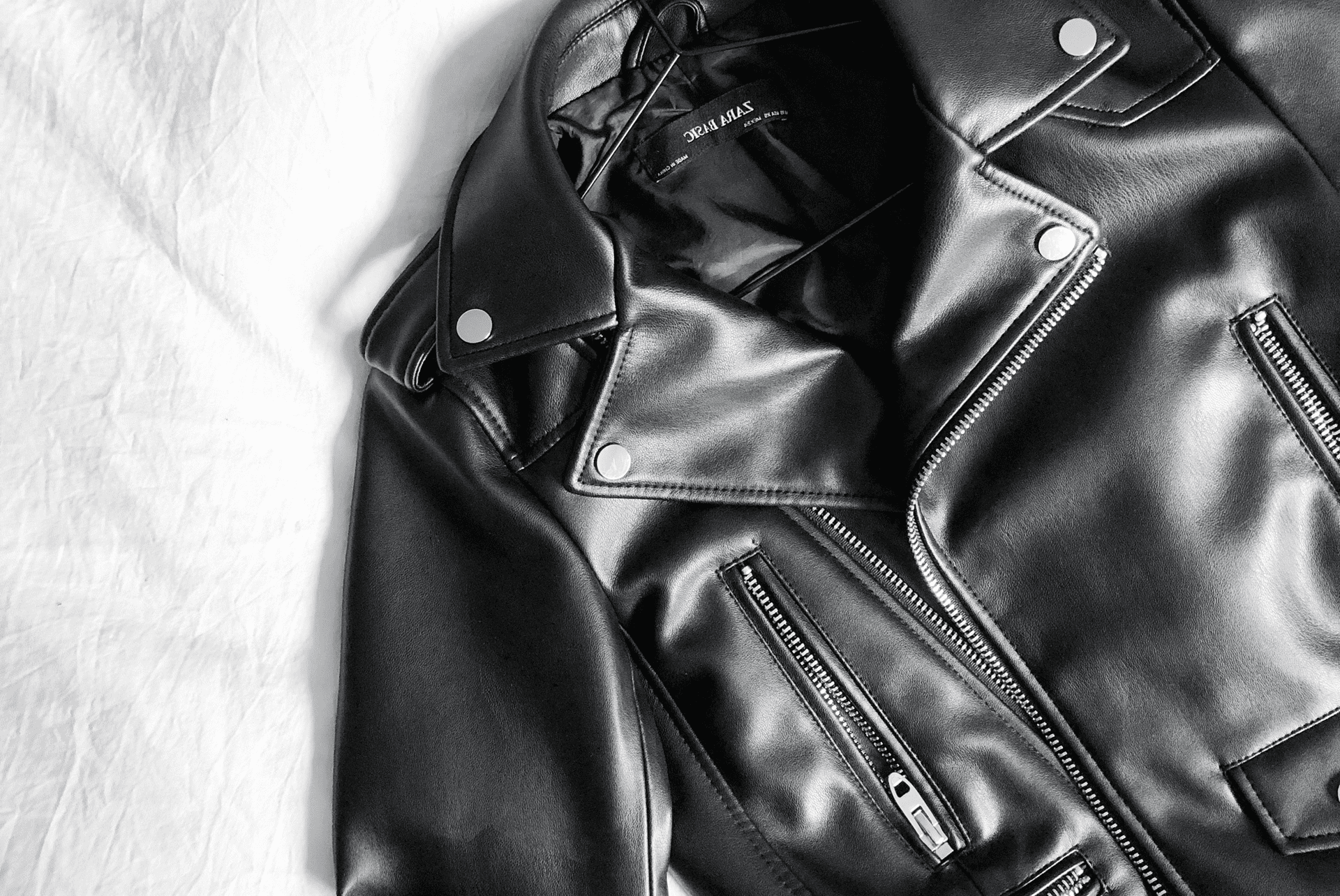 A black leather biker jacket with silver hardware lies on a white fabric background.