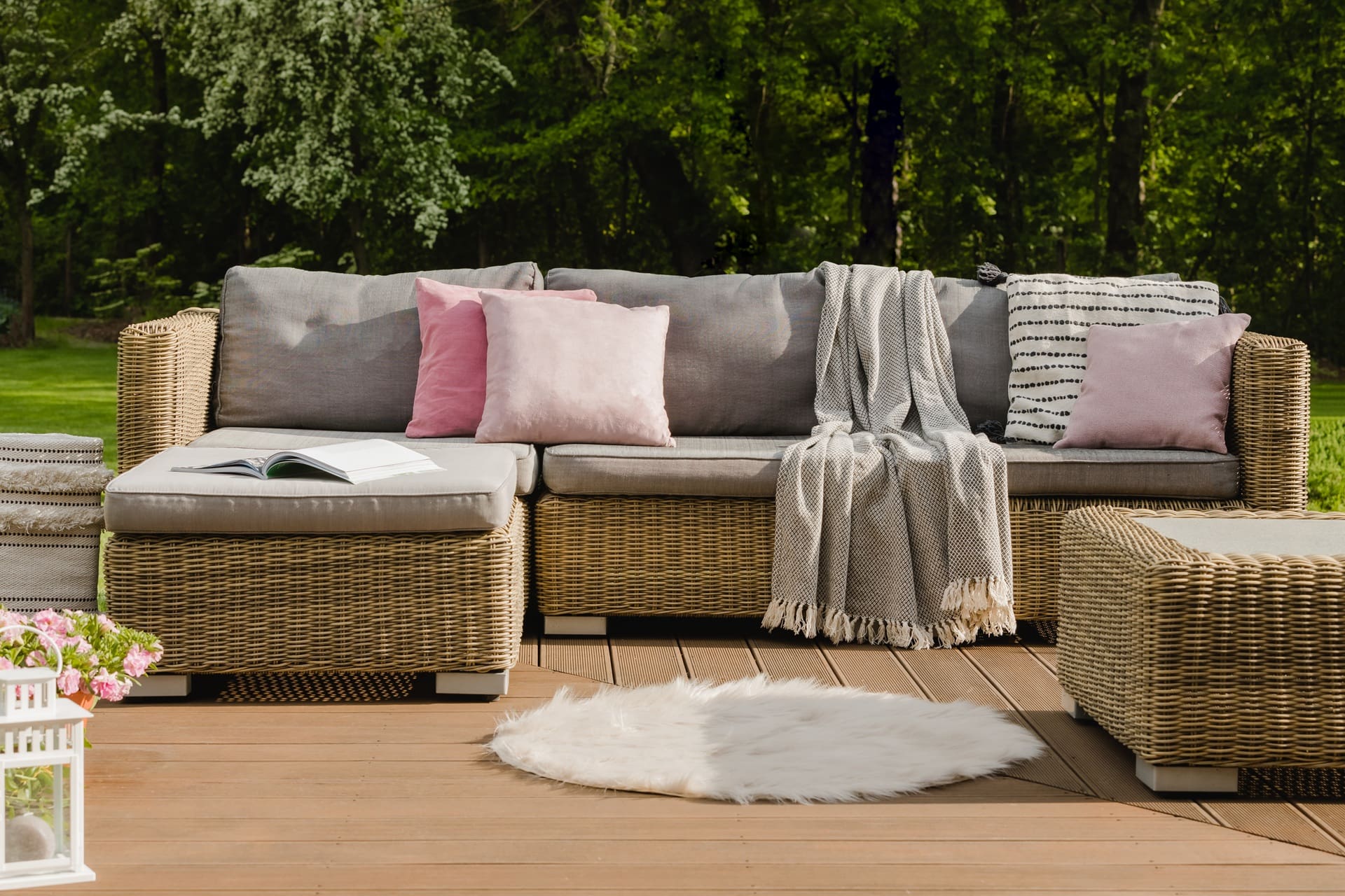 Outdoor wicker sectional sofa with gray cushions, pink and patterned pillows, a draped blanket, and an open book on a wooden deck with green trees in the background.