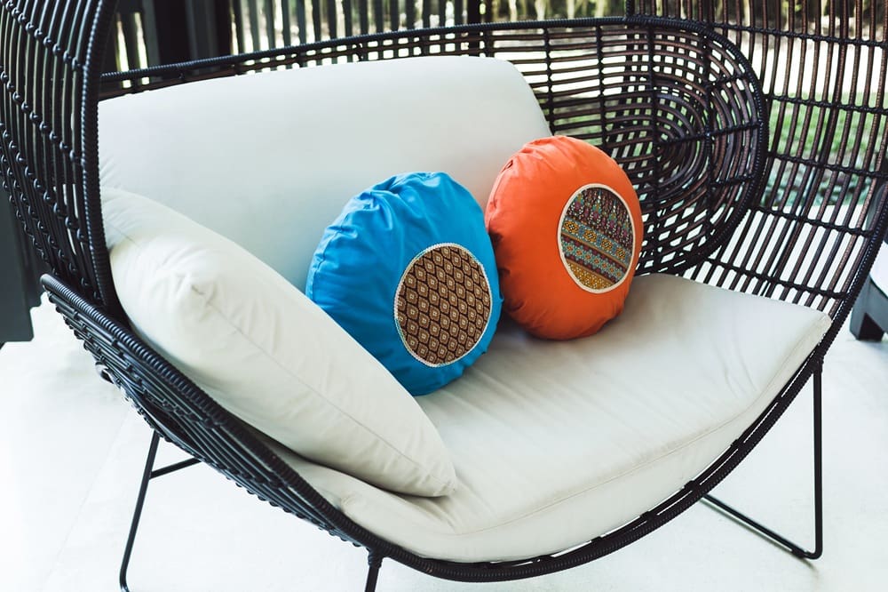 Black wicker loveseat with white cushions and two round pillows, one blue and one orange, each with a patterned center. The loveseat is on a light-colored floor near a window.