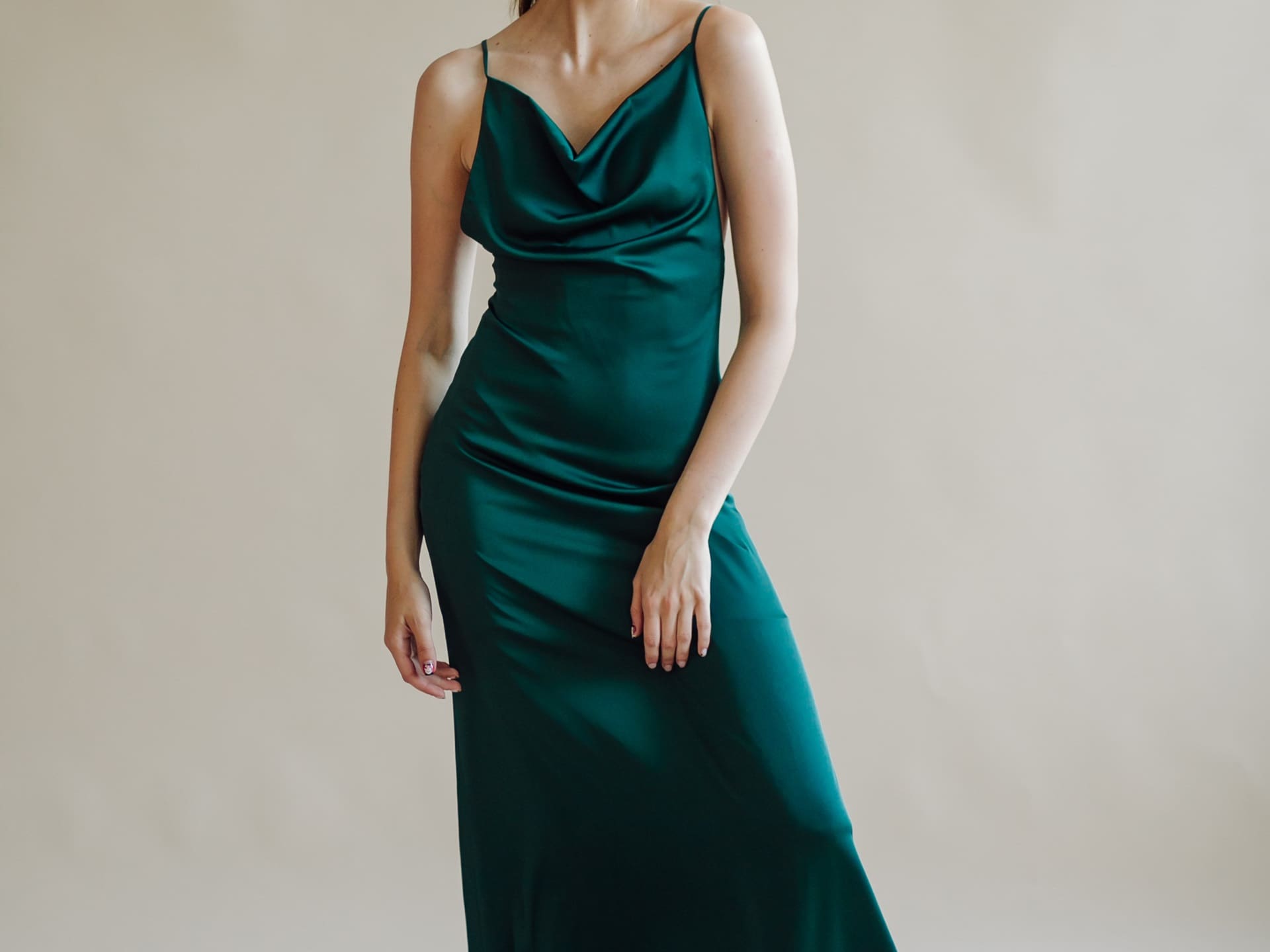 A woman wearing a sleeveless dark green satin dress stands against a plain beige background.