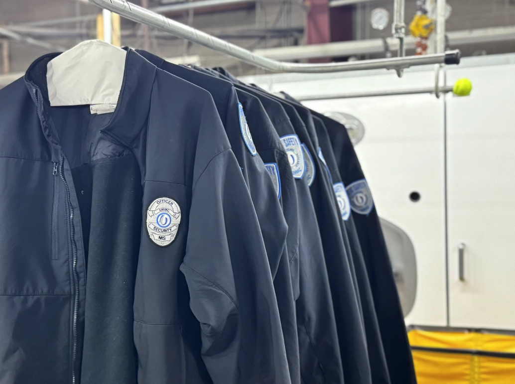 Several identical dark jackets with embroidered patches hang on hangers in a row, inside an industrial or laundry facility.