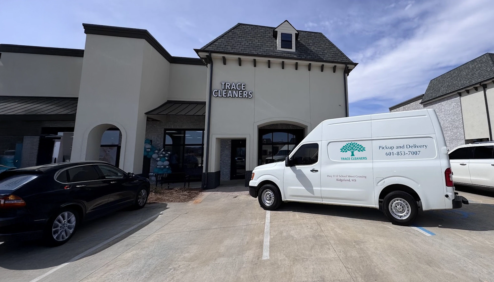 A white delivery van is parked outside Trace Cleaners, a dry cleaning business in a small shopping center, next to a black car under a clear sky.