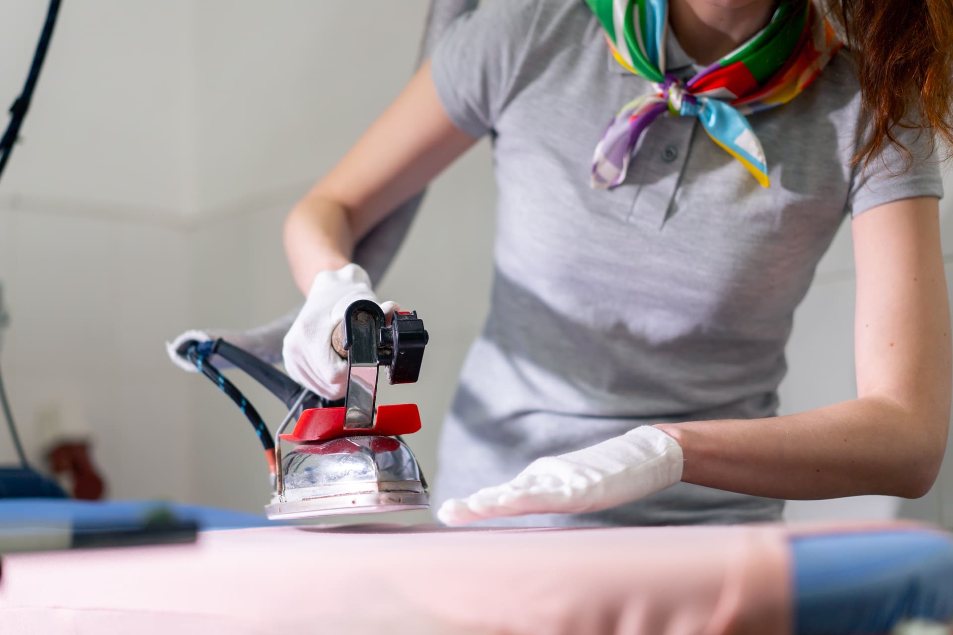Person wearing gloves and a colorful scarf uses a steam iron to press fabric on an ironing board.