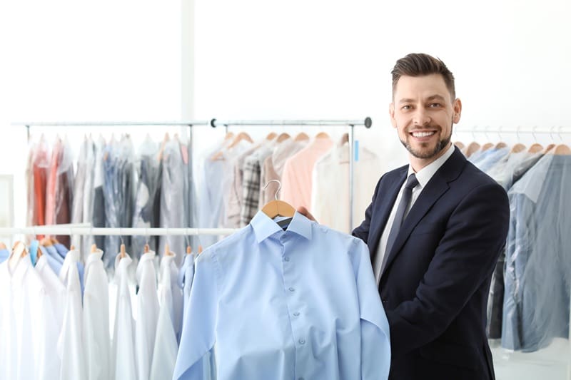 A man in a suit stands in a dry cleaning shop, holding a freshly cleaned blue dress shirt on a hanger. Racks of clothes hang in the background.