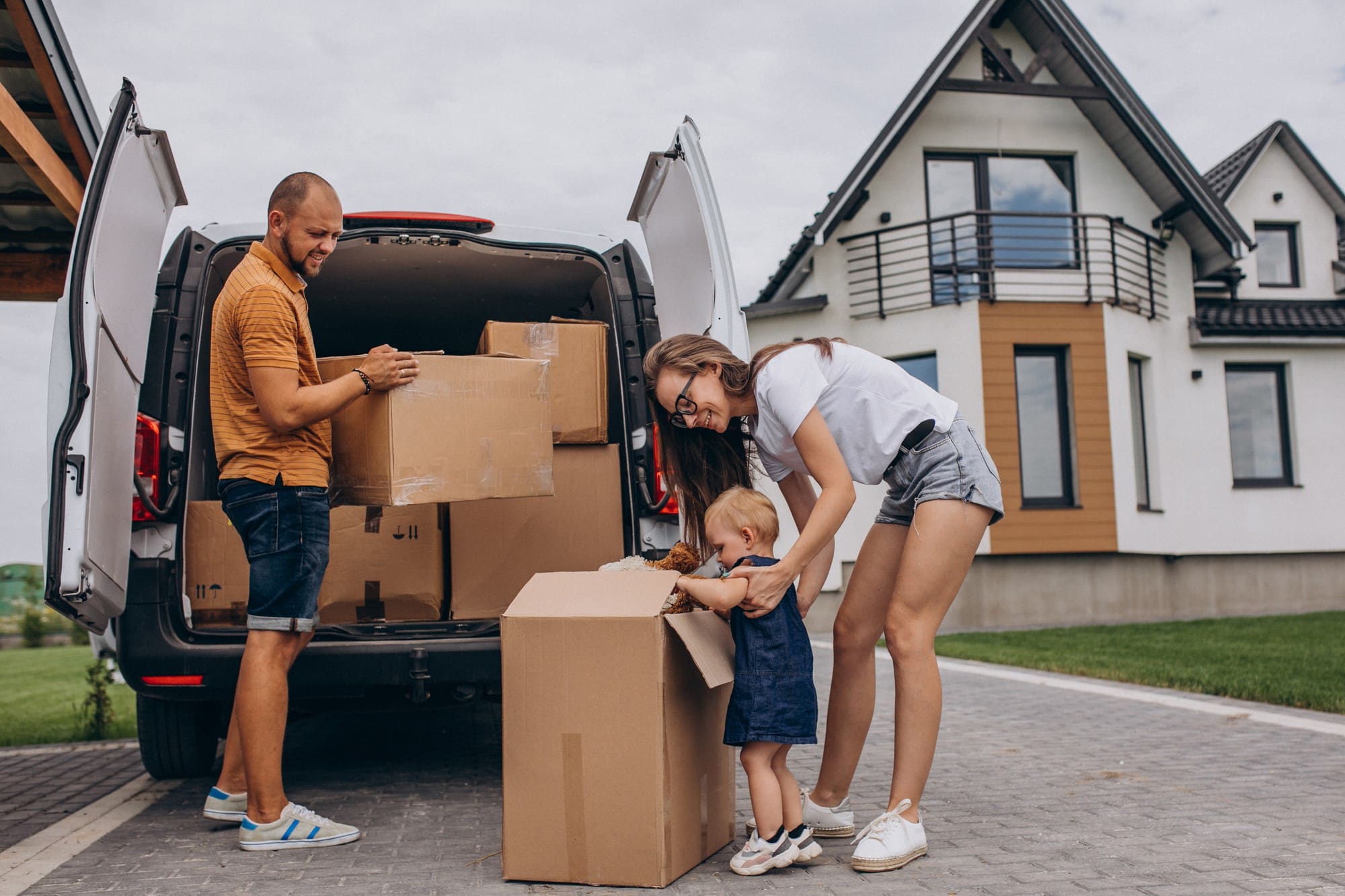 A family unloads cardboard boxes from a van in the driveway of a modern house, with a young child helping beside two adults.