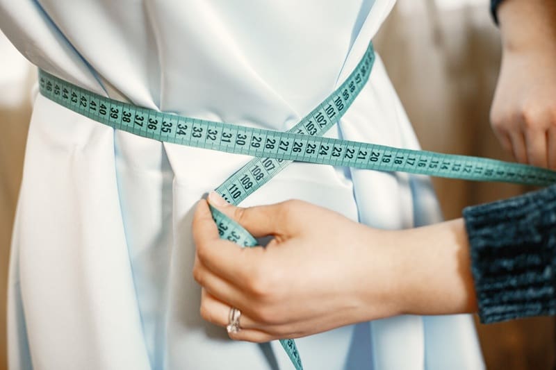 A person wraps a blue measuring tape around the waist of a mannequin dressed in a white garment.