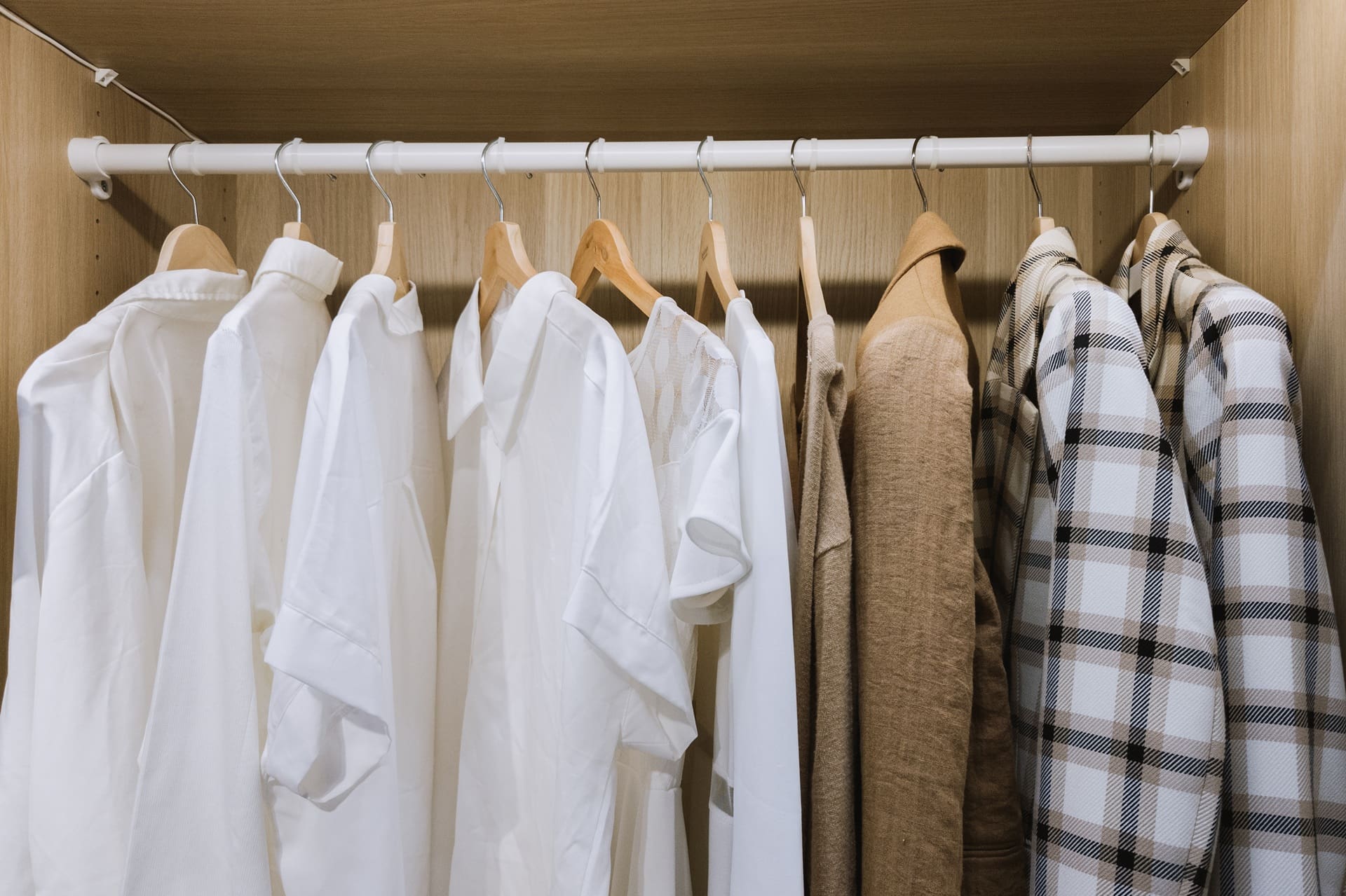 A wardrobe with shirts and jackets in neutral tones, including white, beige, and plaid, neatly hung on wooden hangers on a clothing rod.