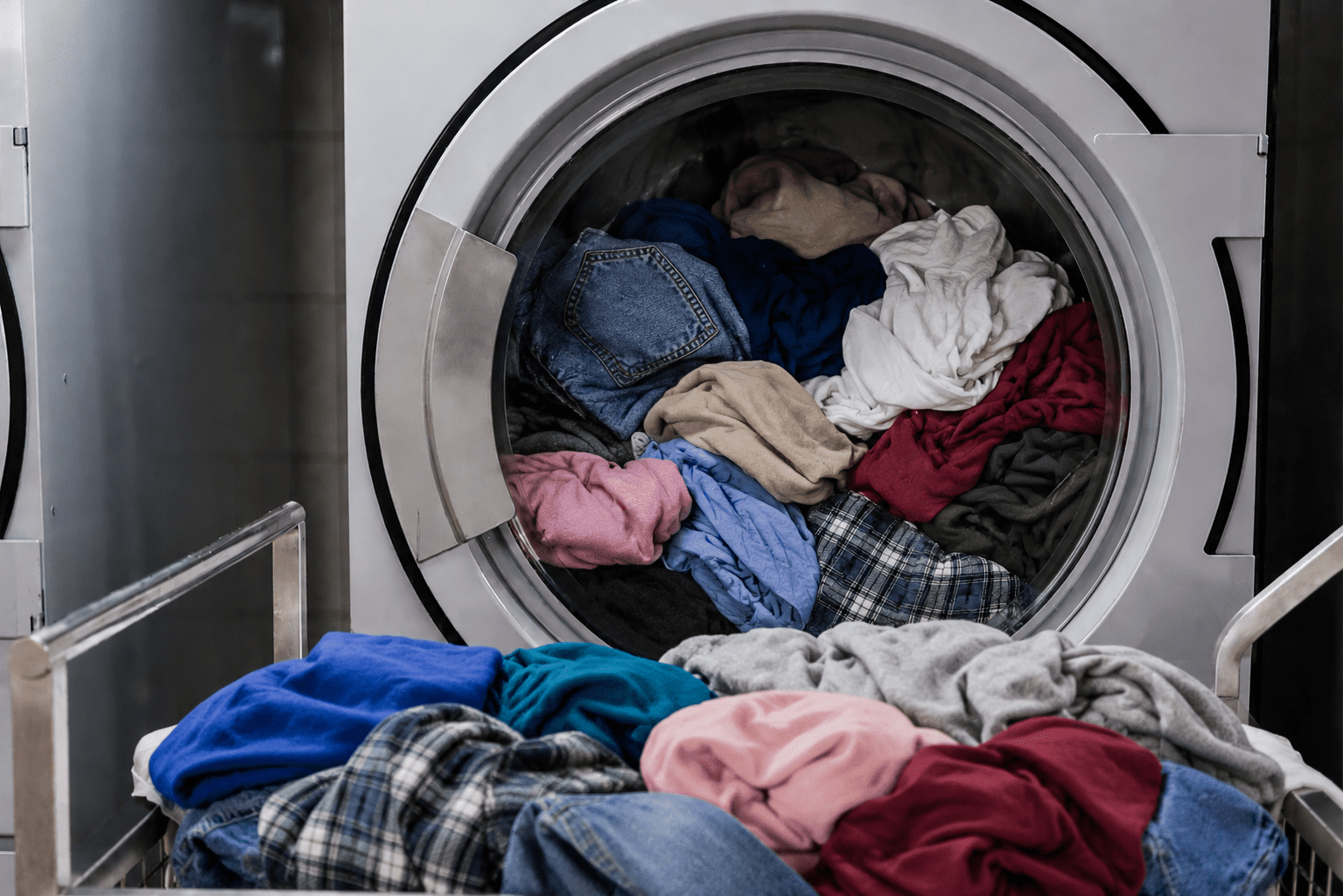 A front-loading washing machine filled with a variety of colorful, mixed laundry, with more clothes piled on the open door and nearby surface.