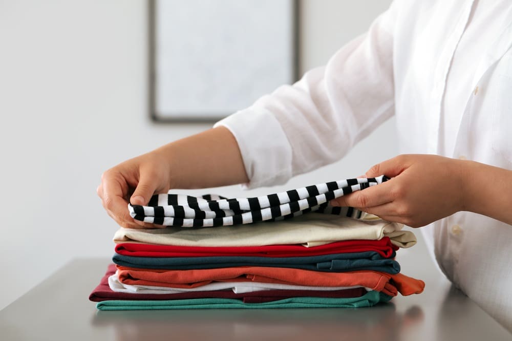 Person folding a stack of neatly arranged clothes, holding a black and white striped shirt on top of several colorful garments.