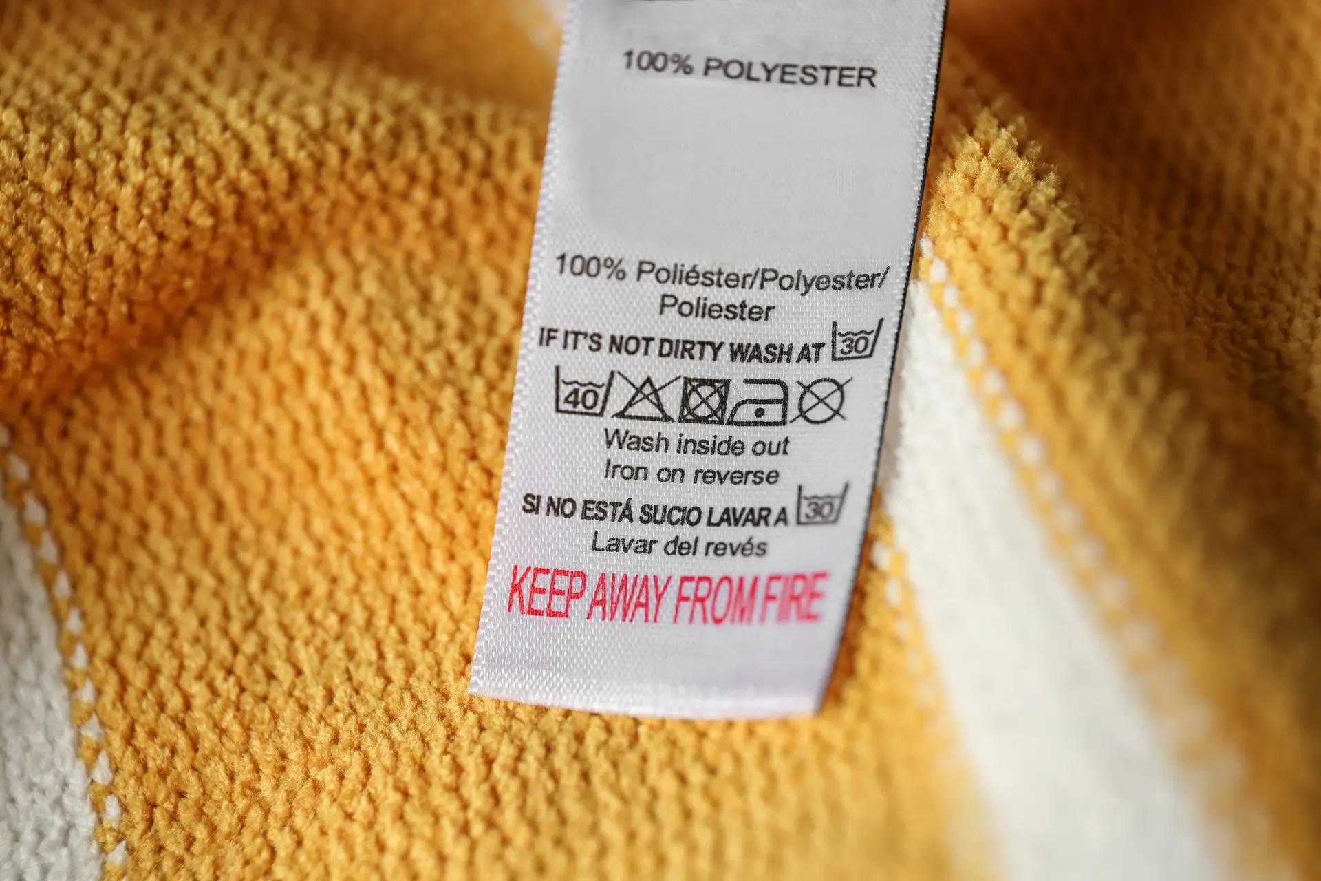 Close-up of a yellow fabric with a care label showing washing instructions, fiber content as 100% polyester, and a warning to keep away from fire.