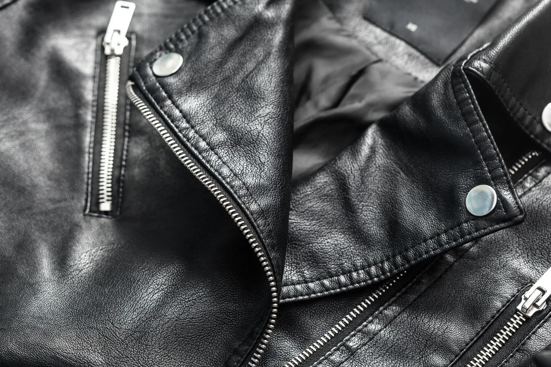 Close-up of a black leather jacket showing silver zippers, snap buttons, and textured material.